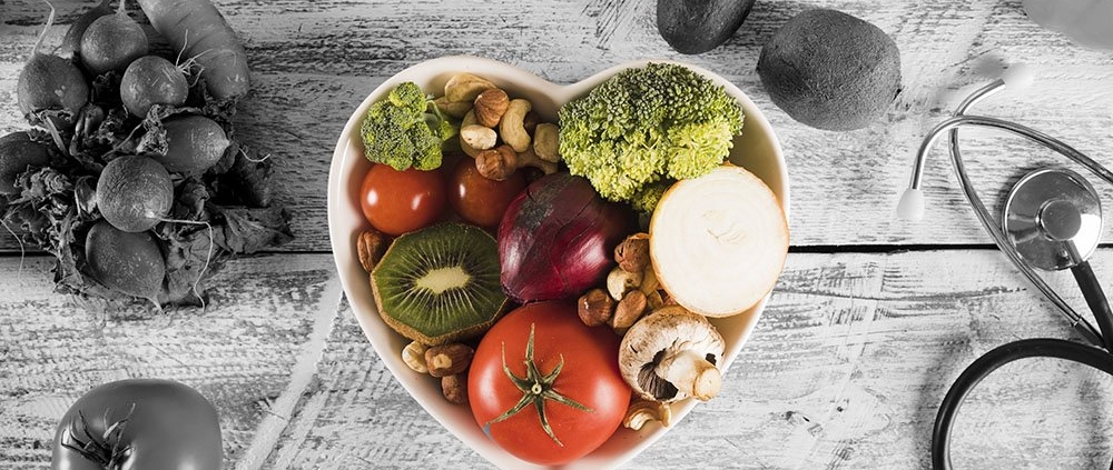 Heart-shaped bowl filled with colorful fruits and vegetables (tomato, kiwi, broccoli, onion, mushrooms, nuts) on a rustic wooden surface, with a stethoscope nearby—symbolizing healthy eating and wellness.