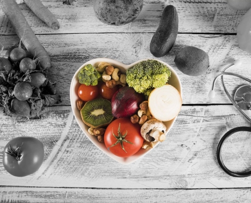 Heart-shaped bowl filled with colorful fruits and vegetables (tomato, kiwi, broccoli, onion, mushrooms, nuts) on a rustic wooden surface, with a stethoscope nearby—symbolizing healthy eating and wellness.