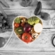Heart-shaped bowl filled with colorful fruits and vegetables (tomato, kiwi, broccoli, onion, mushrooms, nuts) on a rustic wooden surface, with a stethoscope nearby—symbolizing healthy eating and wellness.