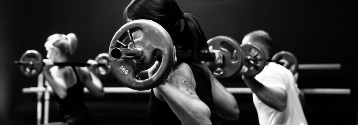 People in a black-and-white gym class performing weighted barbell squats, focus on the central lifter with plate-loaded barbell behind the neck.