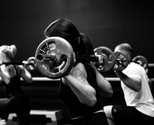 People in a black-and-white gym class performing weighted barbell squats, focus on the central lifter with plate-loaded barbell behind the neck.