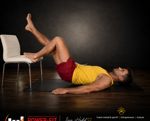 Man lying on his back on a mat with legs raised and resting on a chair, performing a hamstring/hip stretch in a gym setting.