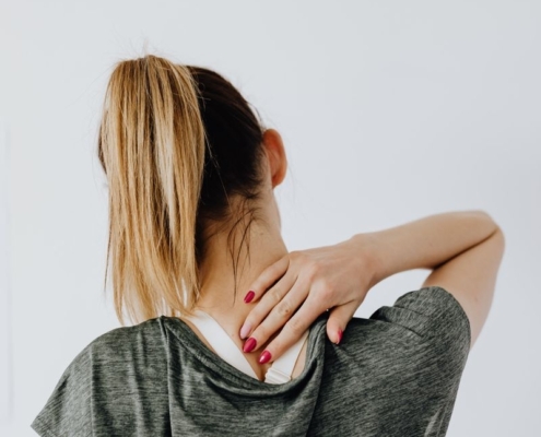 Back view of blond faceless lady in casual t shirt and manicure demonstrating pain spot on back while sitting near white wall