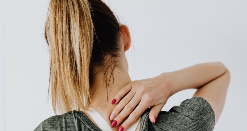Back view of blond faceless lady in casual t shirt and manicure demonstrating pain spot on back while sitting near white wall