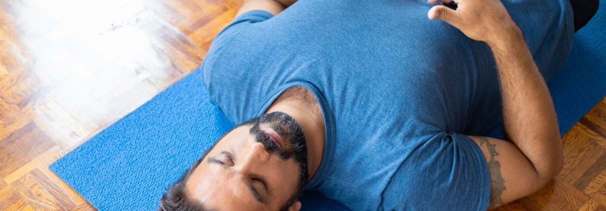 Adult man lying on a blue yoga mat indoors with eyes closed, focusing on relaxation and mindfulness.