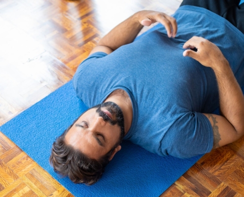 Adult man lying on a blue yoga mat indoors with eyes closed, focusing on relaxation and mindfulness.