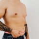 Close-up of a shirtless man measuring his waist with a pink tape measure in a studio setting.